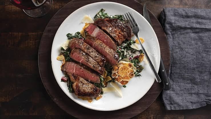Sliced ribeye steak on a white plate with greens and roasted garlic